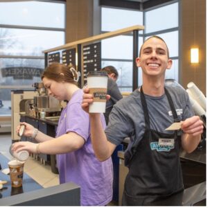 A barista holding a cup and a paper, while another pours a drink behind them.
